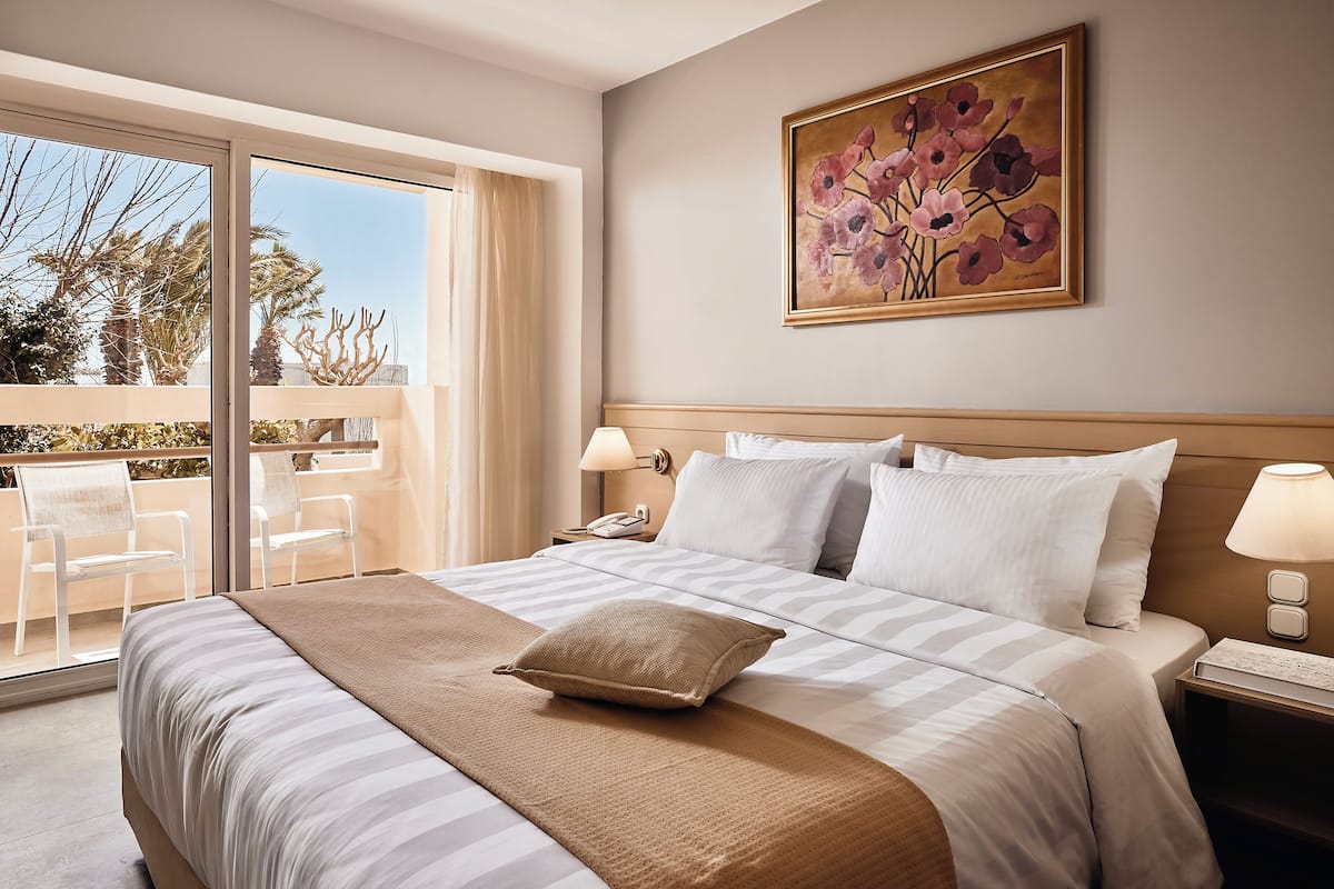 A hotel room with a large bed, a painting on the wall, a balcony with chairs, and a view of palm trees.