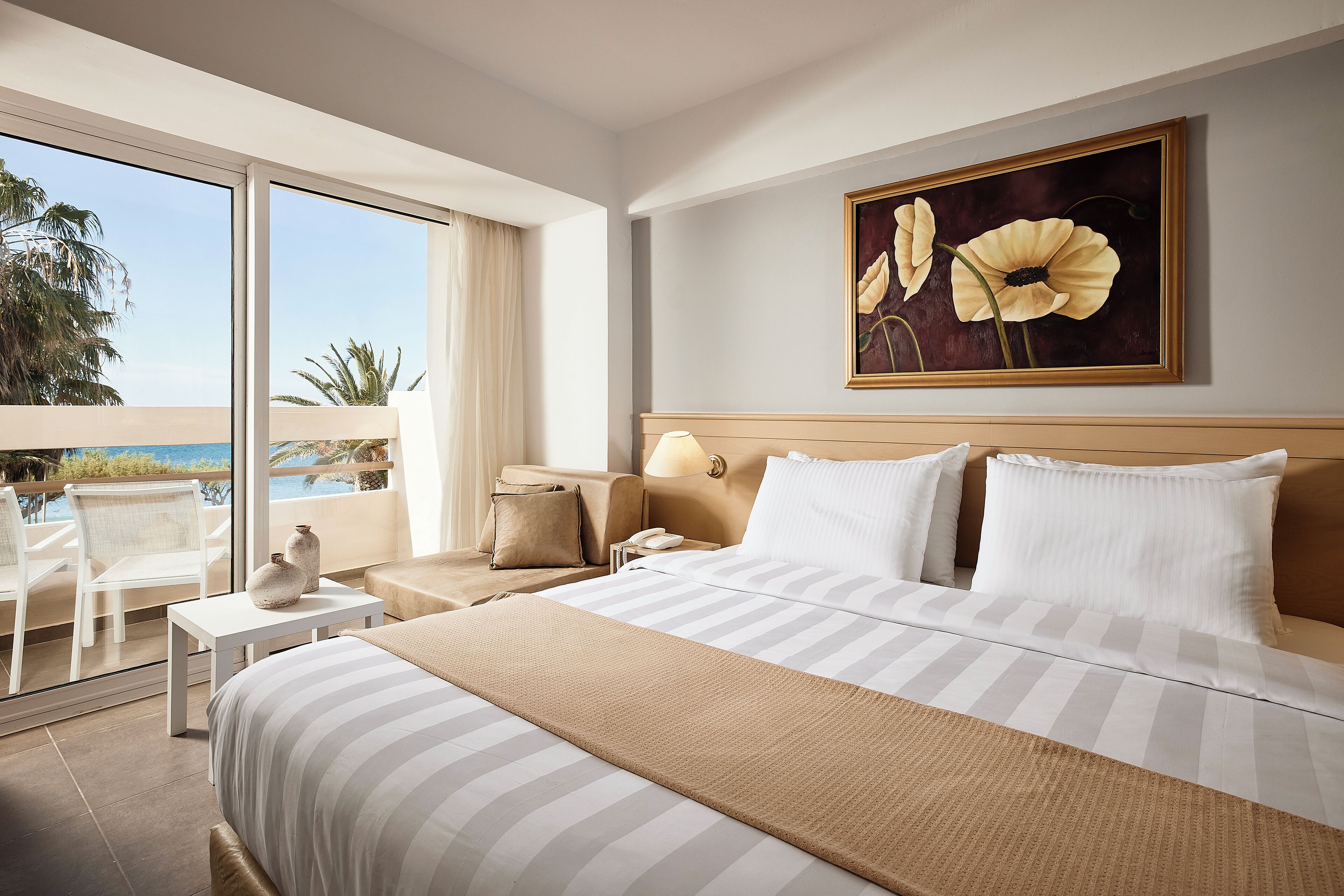 A hotel room with a large bed, a painting of flowers on the wall, a balcony with a view of the ocean, and a small table with a vase.