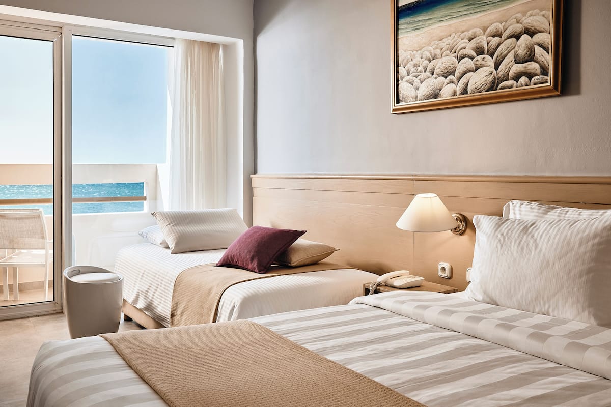 A hotel room with two beds, a view of the sea, a painting on the wall, and a lamp on the nightstand.
