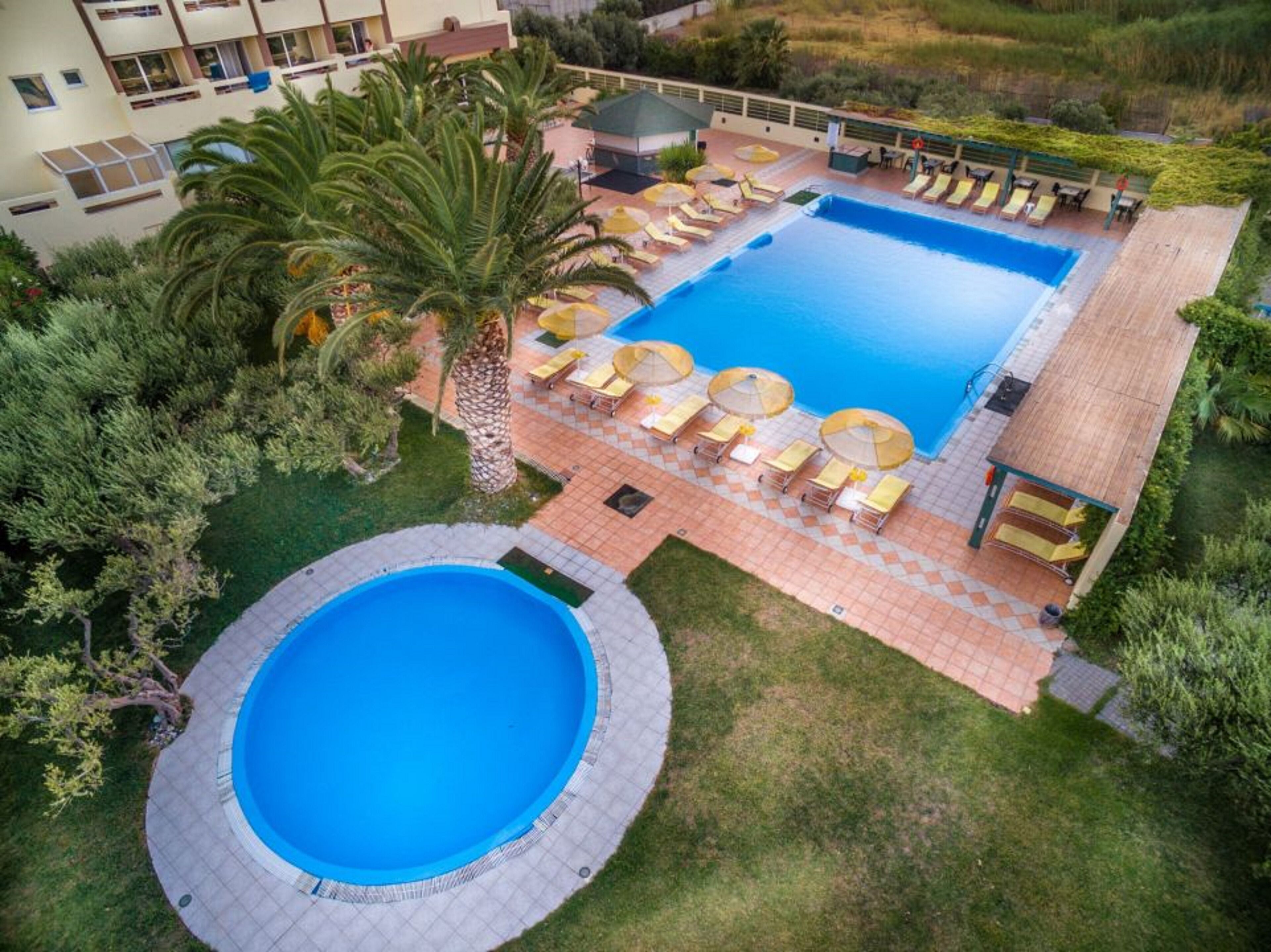 A large outdoor swimming pool with a circular section, surrounded by lounge chairs and umbrellas, and a small building in the background.