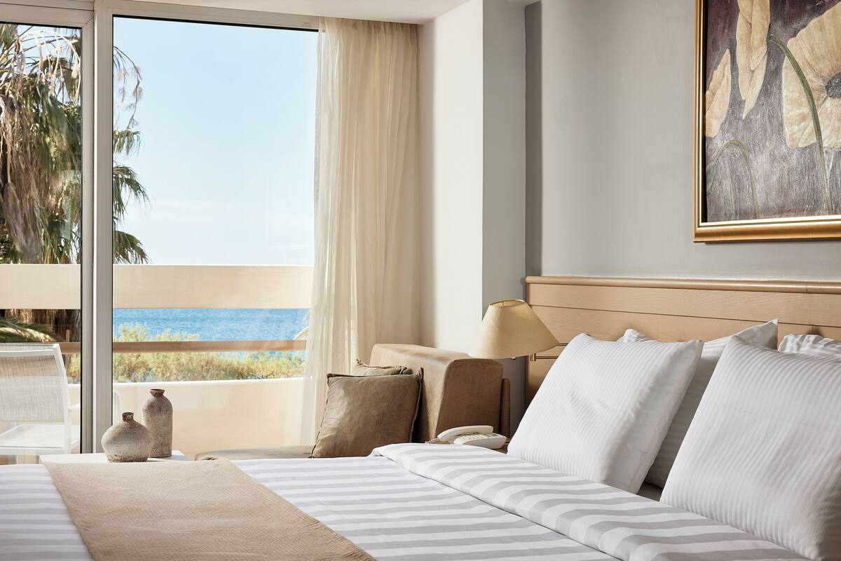 A hotel room with a bed, a bedside table, a lamp, and a view of the ocean.