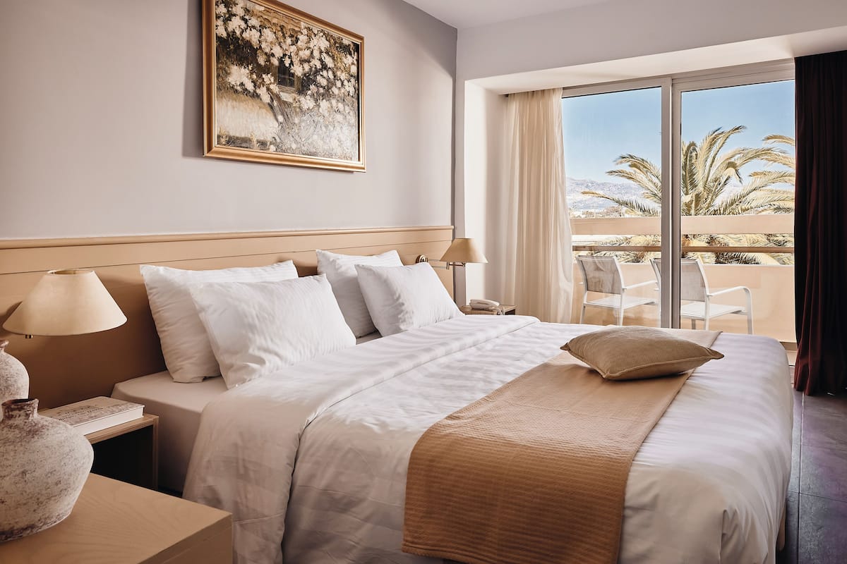 A hotel room with a large bed, a nightstand with a lamp, a painting on the wall, and a view of palm trees through the window.