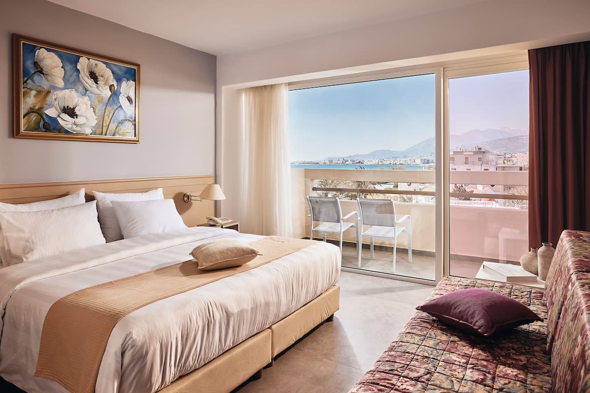 A hotel room with a large bed, a sofa, a balcony with a view of the city and mountains, and a painting on the wall.
