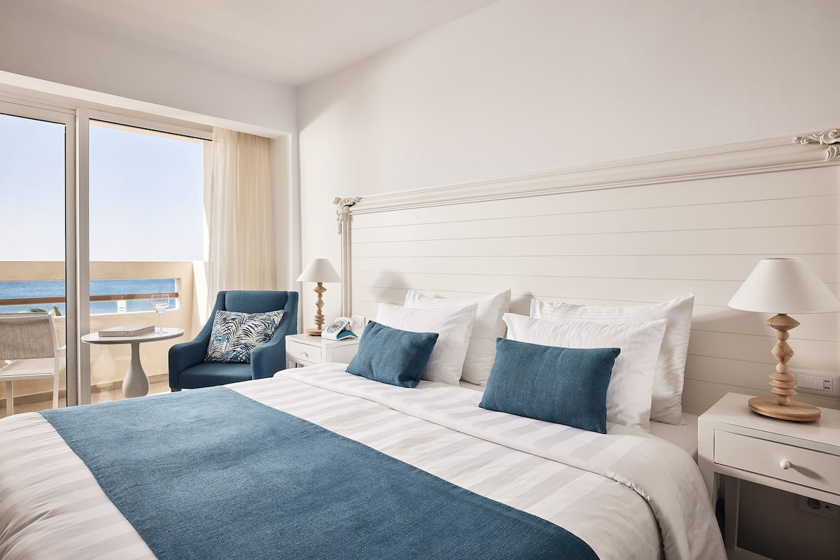 A hotel room with a large bed, a chair, a small table, and a view of the sea.