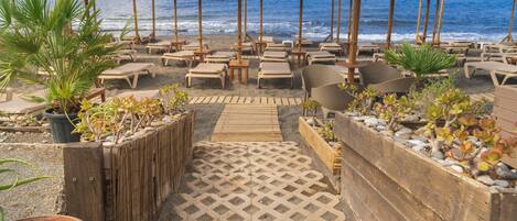 Een strandlounge met rieten parasols, lounge fauteuils en potplanten.