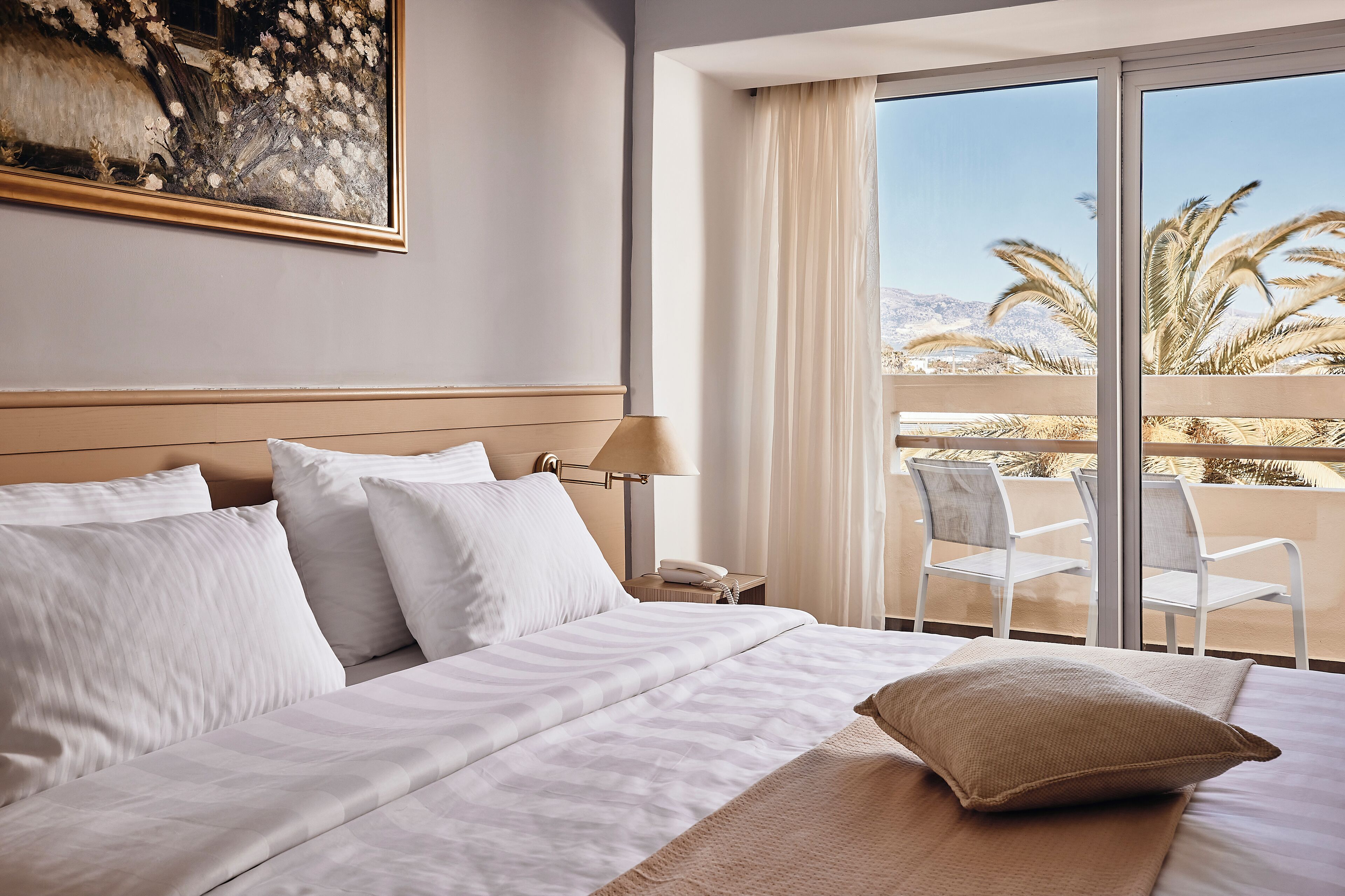 Habitación de hotel con una cama grande, un cuadro en la pared, un balcón con sillas y vistas a palmeras.