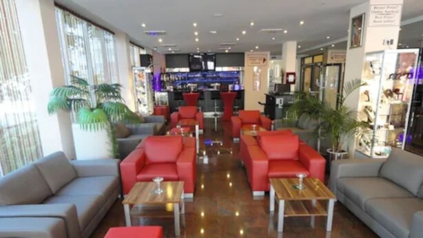 A hotel lobby with red and grey sofas, wooden tables, and a bar area in the background. - Grand Hotel Dream Frankfurt City (Frankfurt)