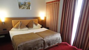A double bed with two pillows and a blanket, a nightstand with a lamp, a painting on the wall, and striped curtains. - Grand Hotel Dream Frankfurt City (Frankfurt)