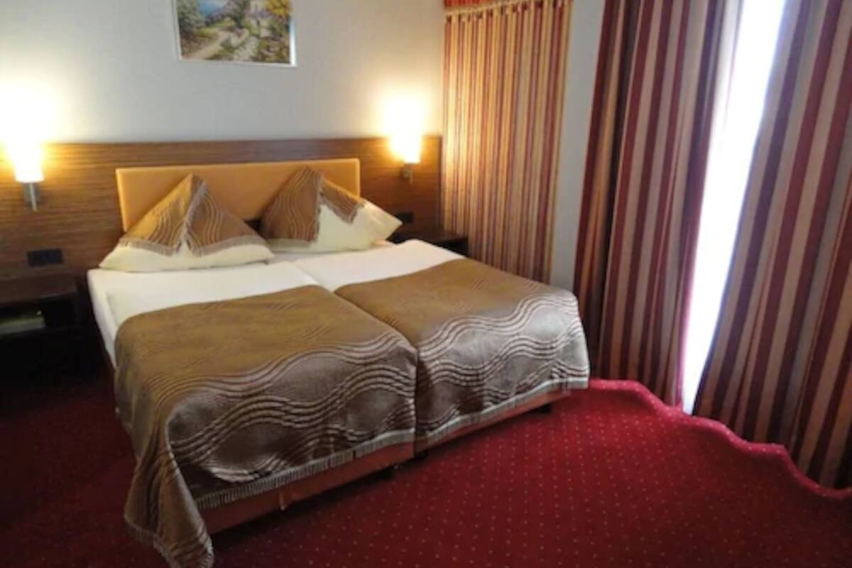 A double bed with two pillows and a blanket, a nightstand with a lamp, a painting on the wall, and striped curtains.