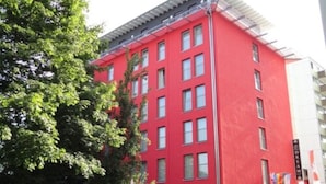 A red building with multiple windows and a flat roof, surrounded by trees and parked cars. - Grand Hotel Dream Frankfurt City (Frankfurt)