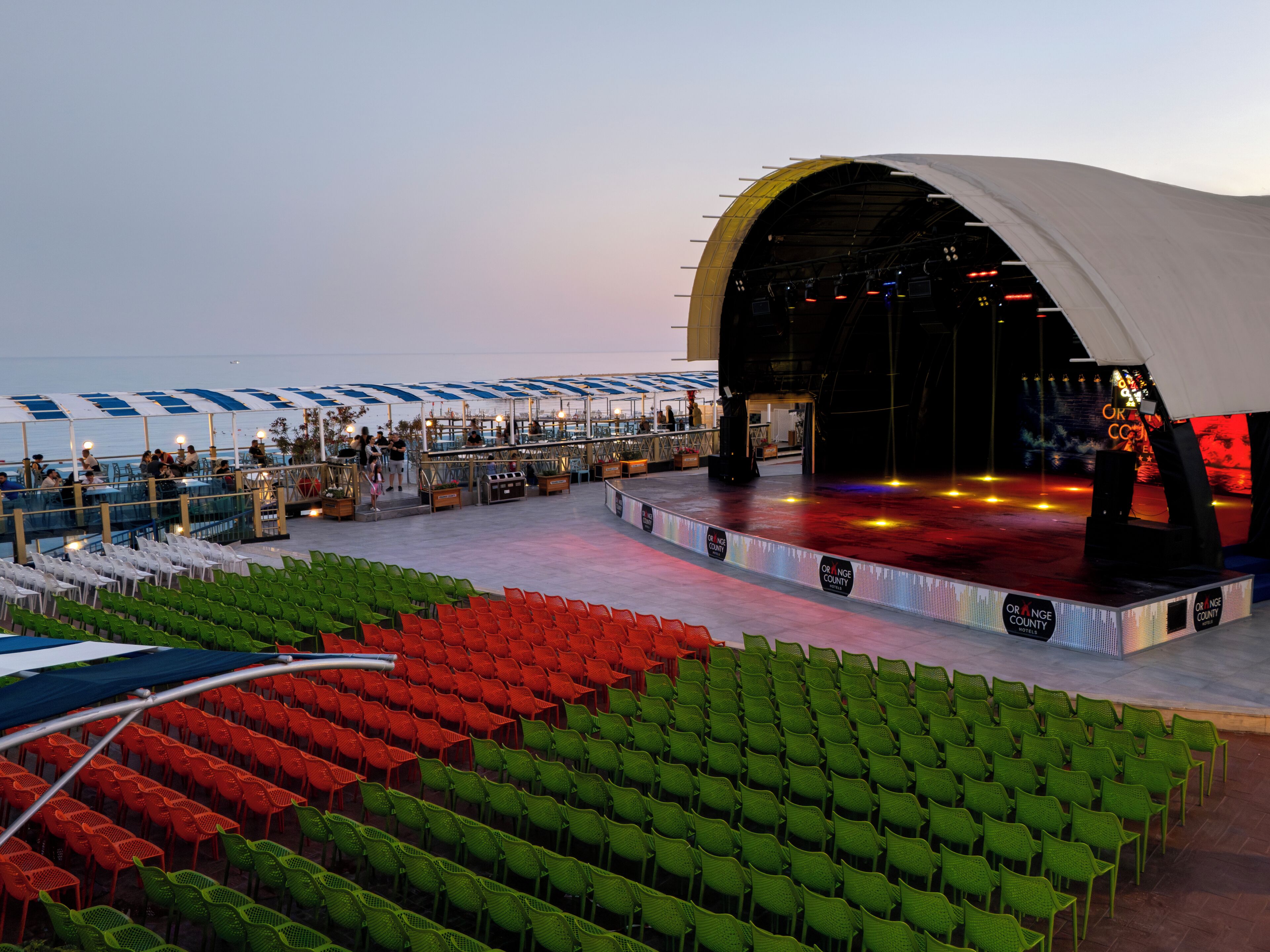 A stage with a curved roof, seating areas with green and red chairs, and a view of the ocean.