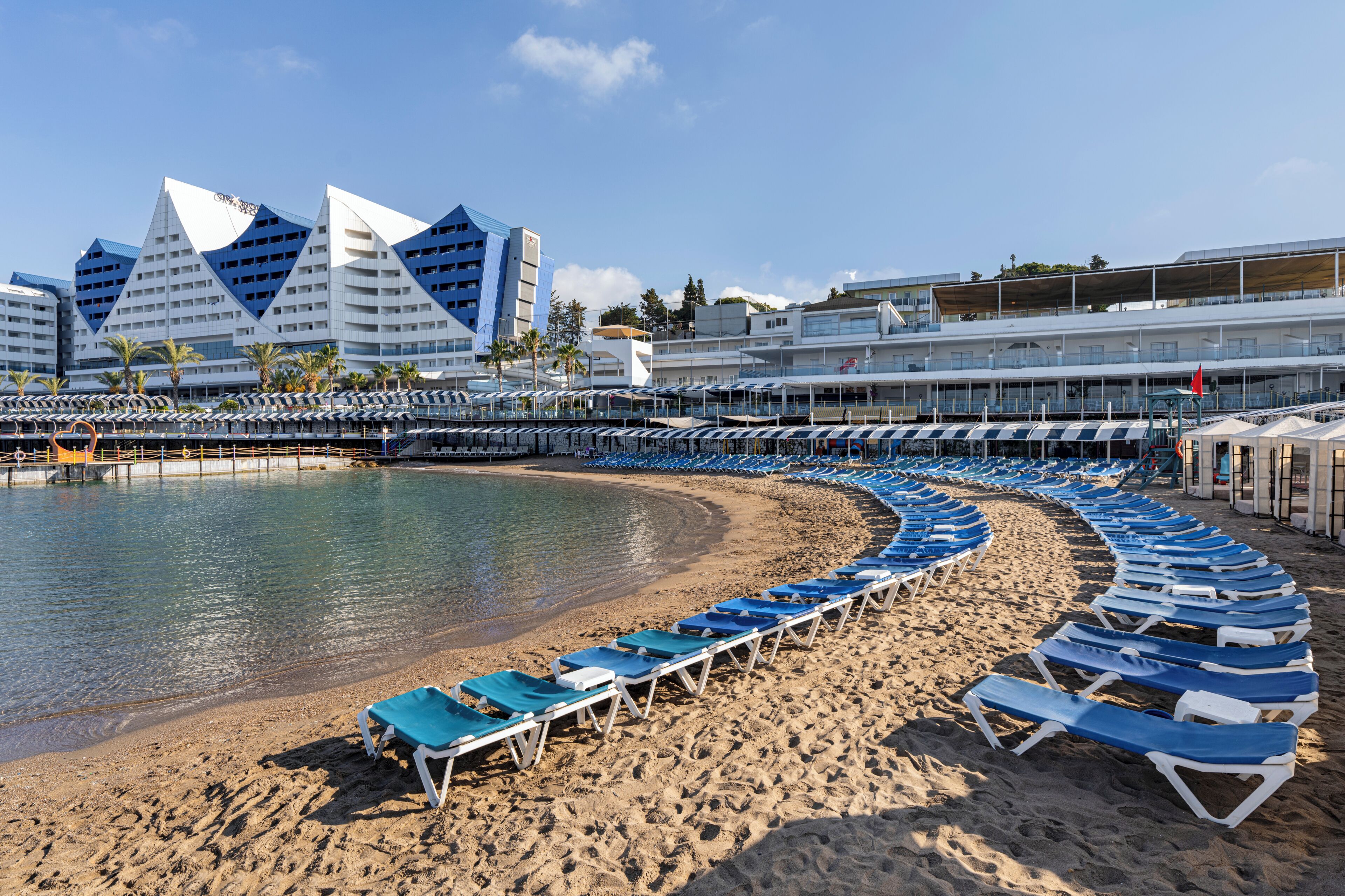 A beachside hotel with a curved row of sun loungers, a pool, and modern buildings in the background.