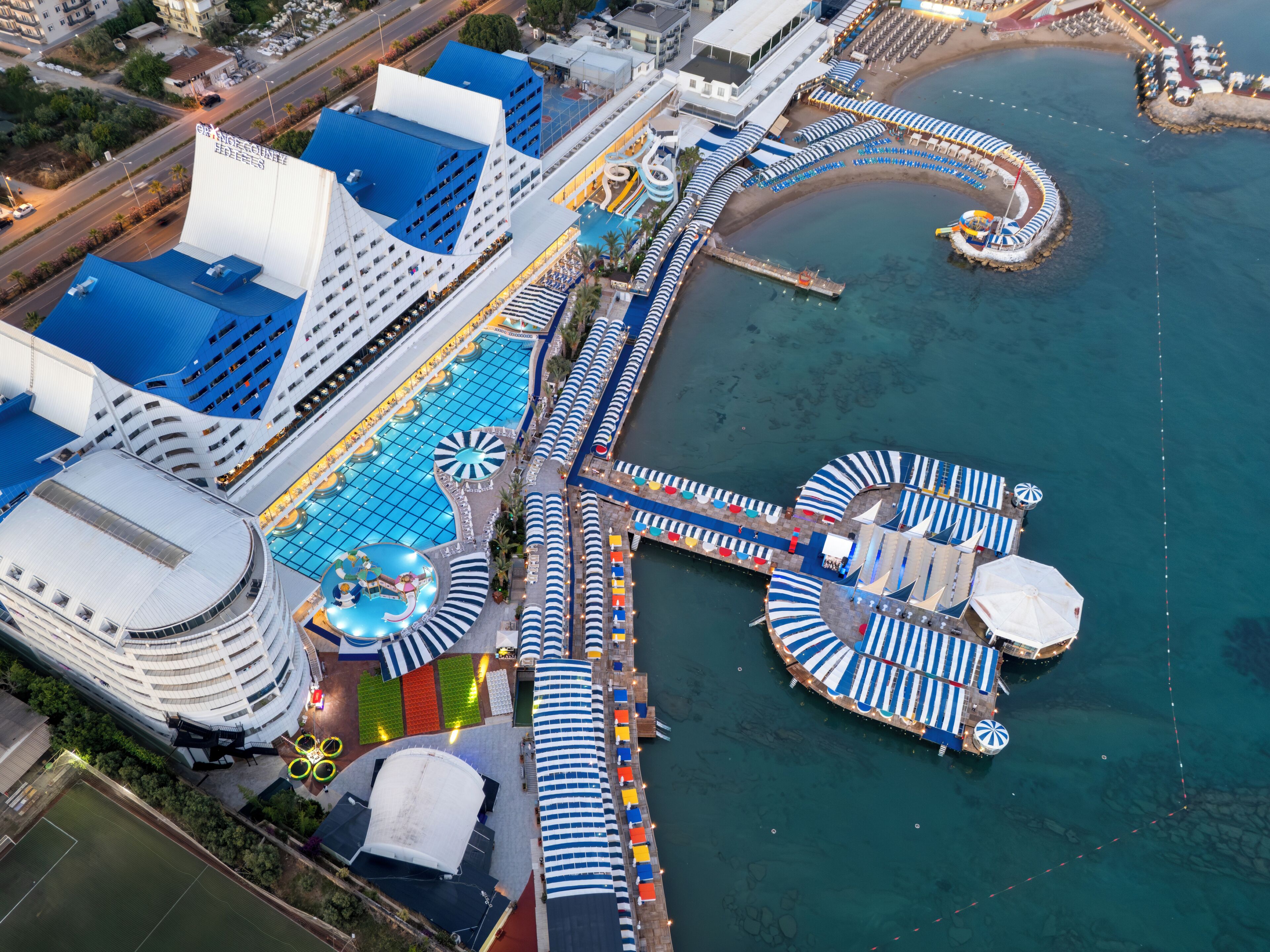 A cruise ship docked at a port with a swimming pool, sun loungers, and a pier extending into the water.