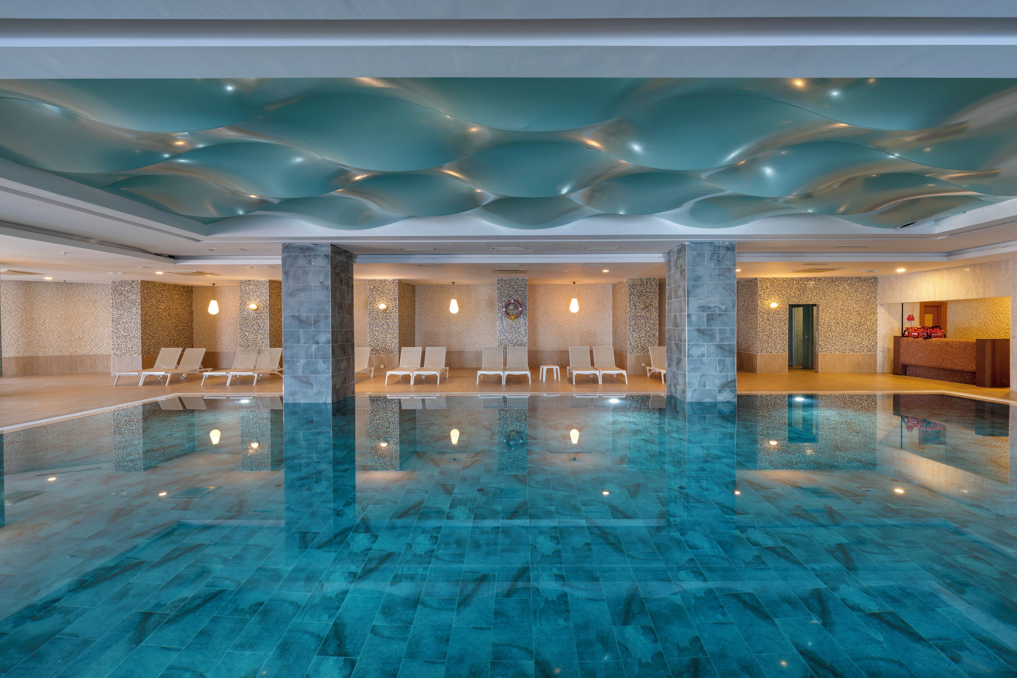 Ein großzügiges Hallenbad mit klarem Wasser, Liegestühlen und einem modernen Deckenbau.