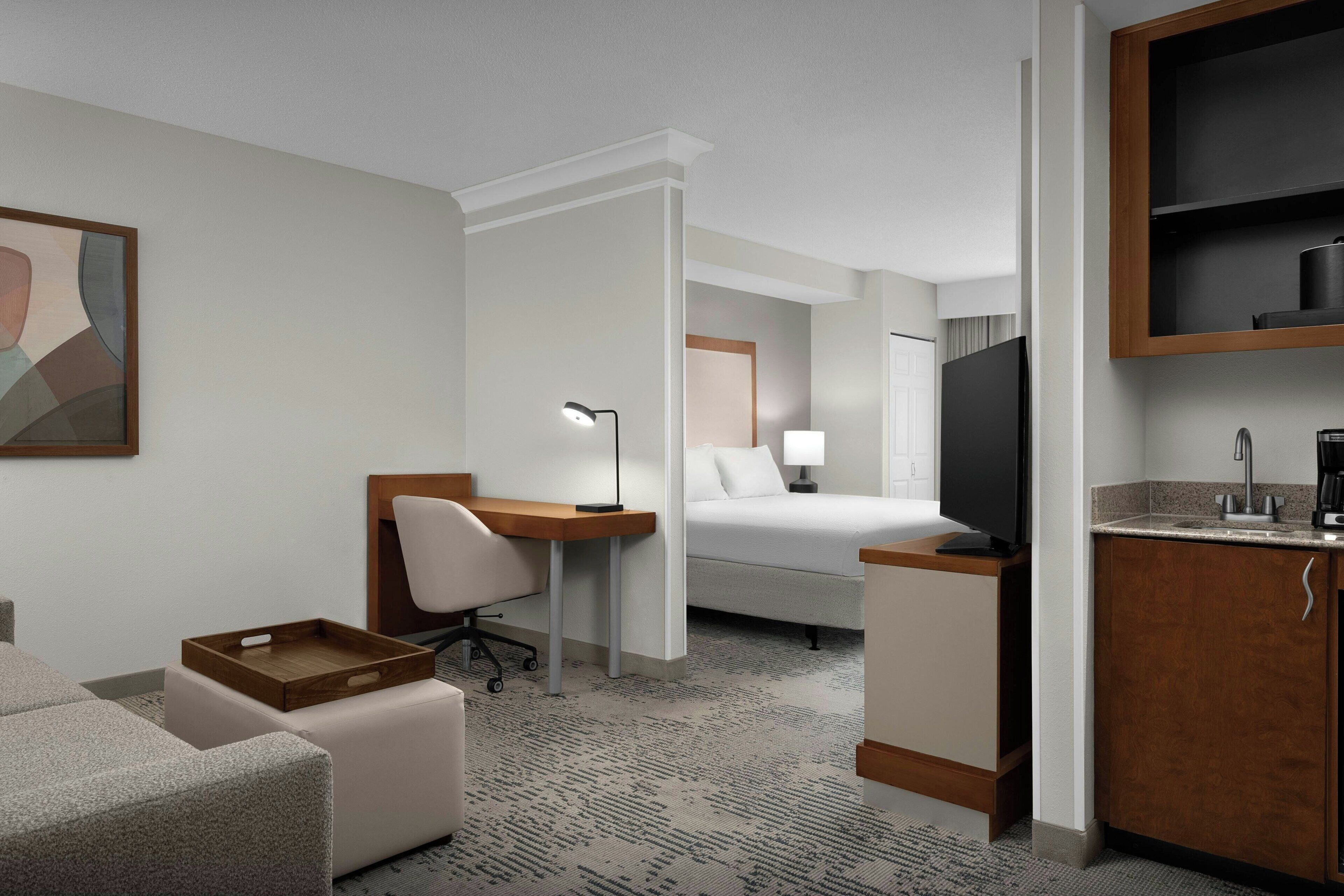 A hotel room with a bed, desk, chair, TV, and kitchenette.