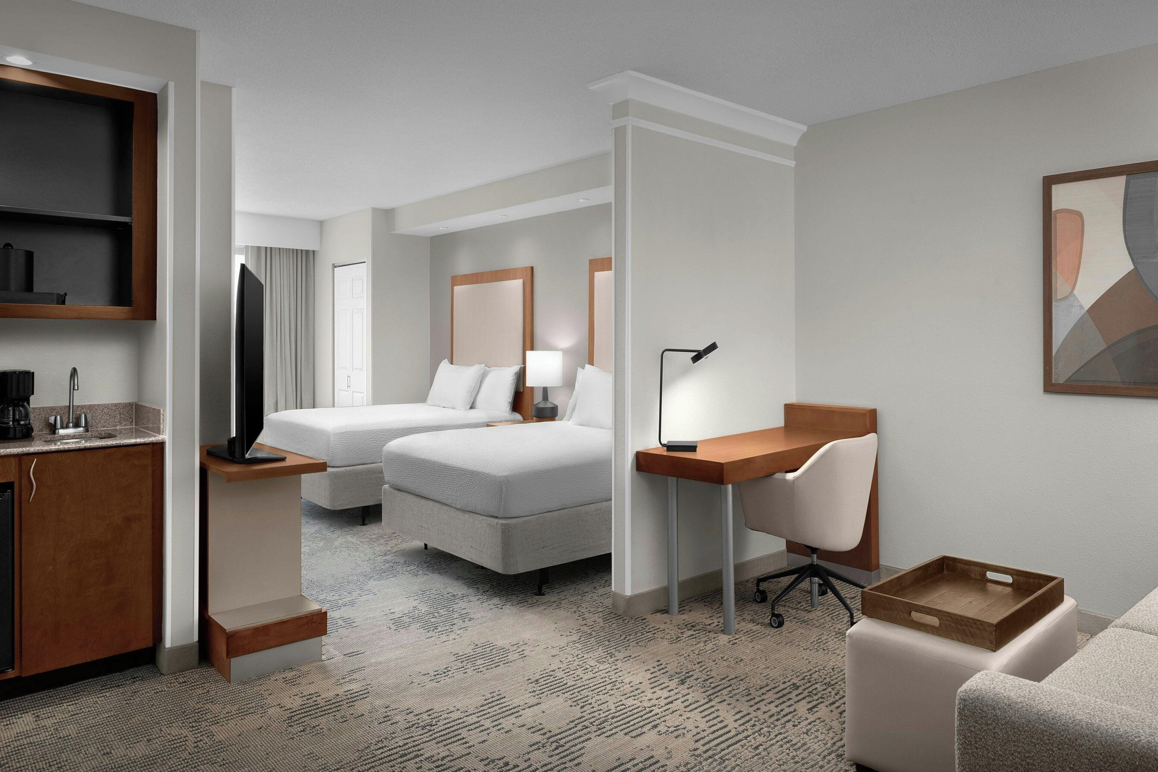 Quarto de hotel com cama, escrivaninha, cadeira, televisão e uma pequena cozinha.