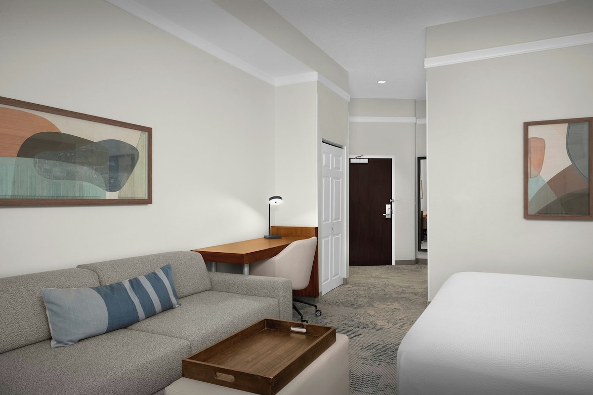 Ett hotellrum med en grå soffa, ett skrivbord i trä, en vit säng och abstrakt konst på väggarna.