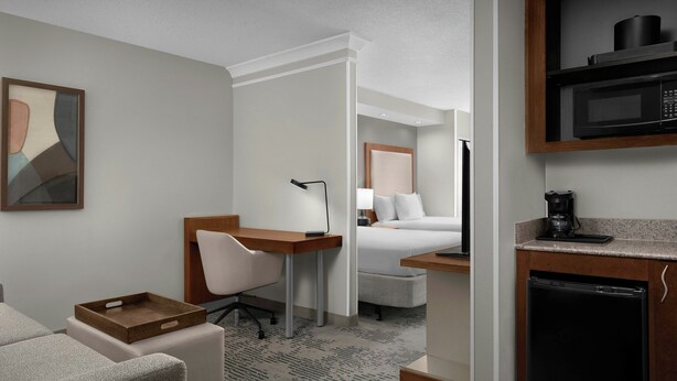 Habitación de hotel con cama, escritorio con silla, microondas, cafetera y refrigerador pequeño.