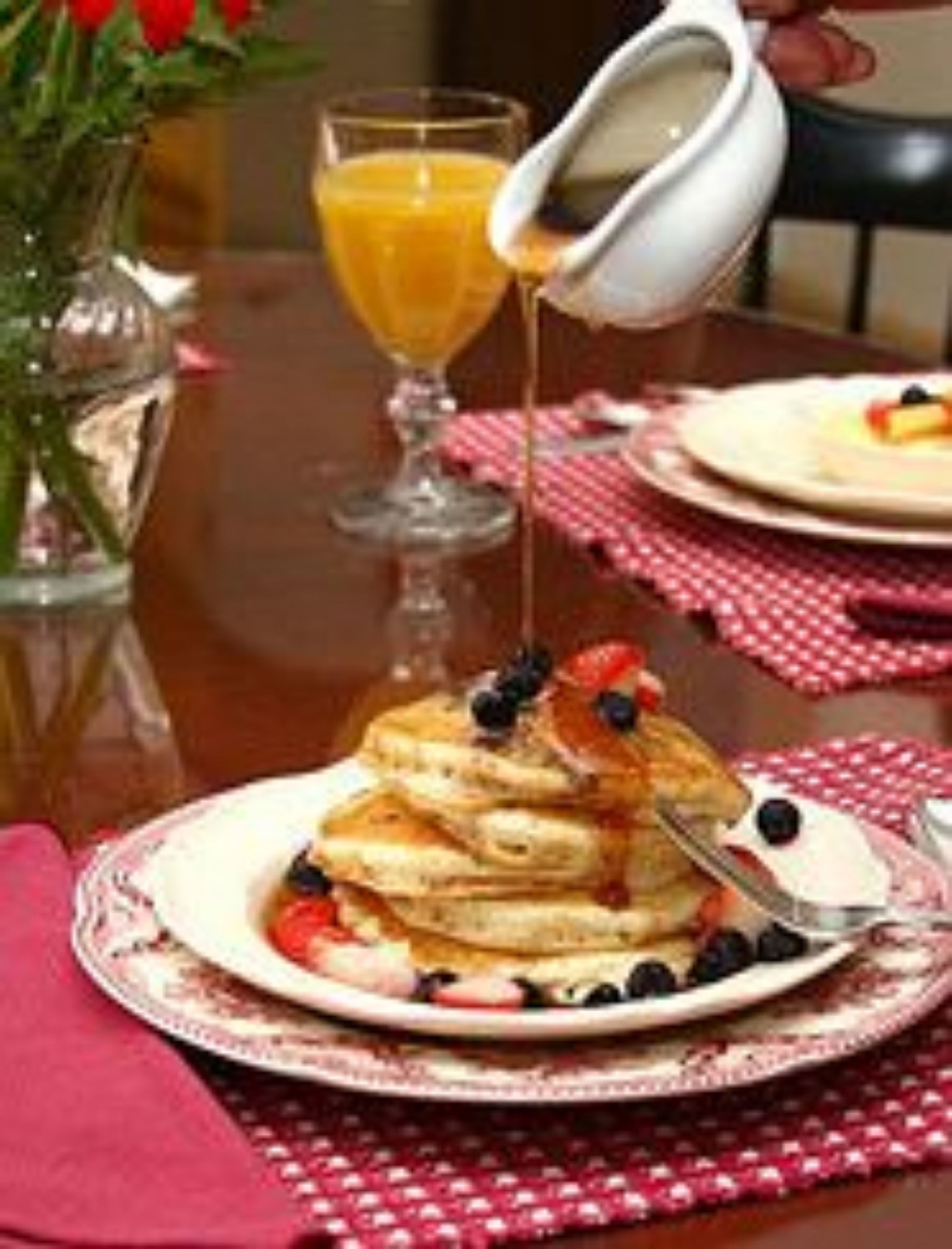 A breakfast setting with pancakes topped with blueberries and syrup, a glass of orange juice, and a pitcher of syrup on a table with a red and white checkered tablecloth.