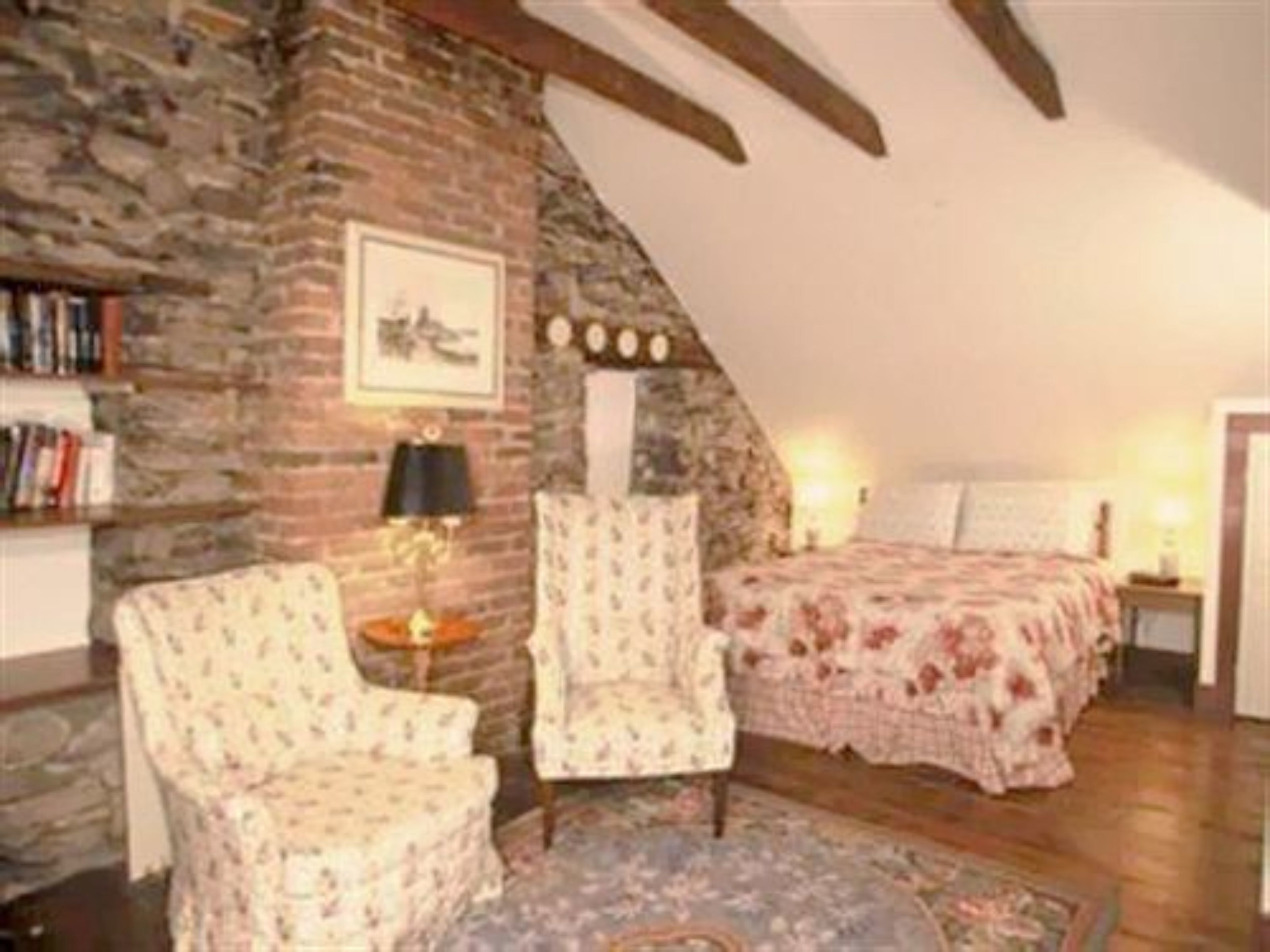 A cozy room with a sloped ceiling, a brick wall, a bed with floral bedding, two armchairs, a small table, and a bookshelf.