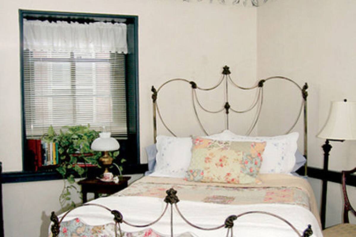 Quarto com cama de dossel, janela com persianas, mesa de cabeceira com abajur e criado-mudo com luminária.
