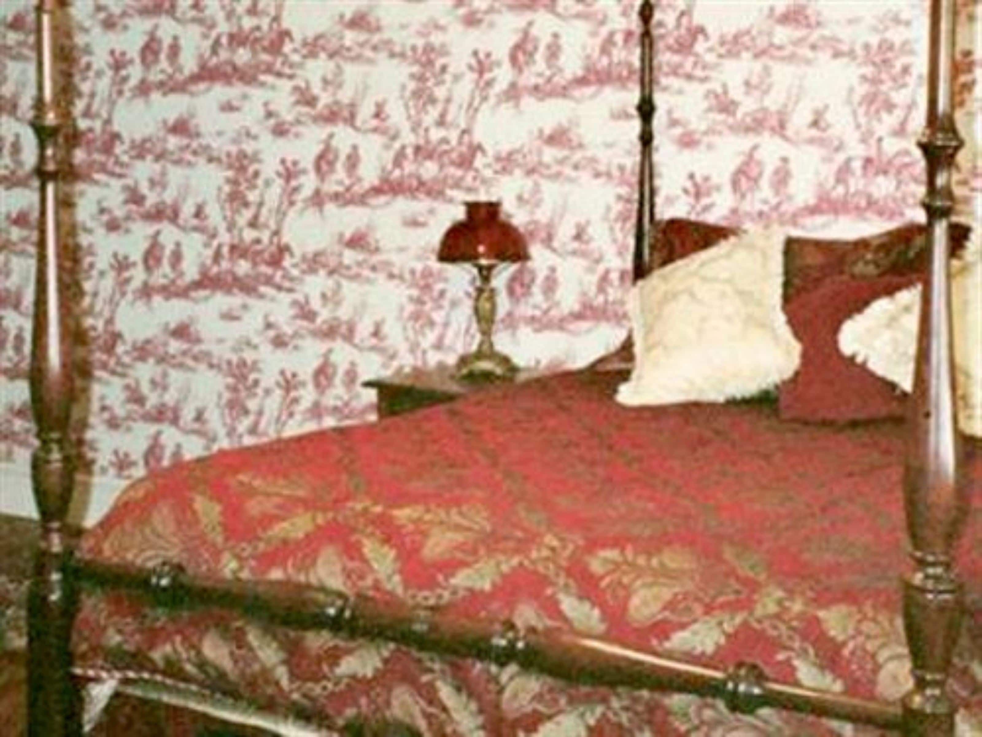 Un lit à baldaquin avec une couvre-lit aux motifs rouges et dorés, un oreiller blanc et moelleux, et une lampe rouge posée sur une table de chevet.