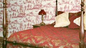 Un lit à baldaquin avec une couvre-lit aux motifs rouges et dorés, un oreiller blanc et moelleux, et une lampe rouge posée sur une table de chevet.
