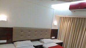 A hotel room with a double bed, a bedside table, a nightstand, a wall-mounted air conditioner, and a window with curtains. - Hotel Milan International (Mumbai)