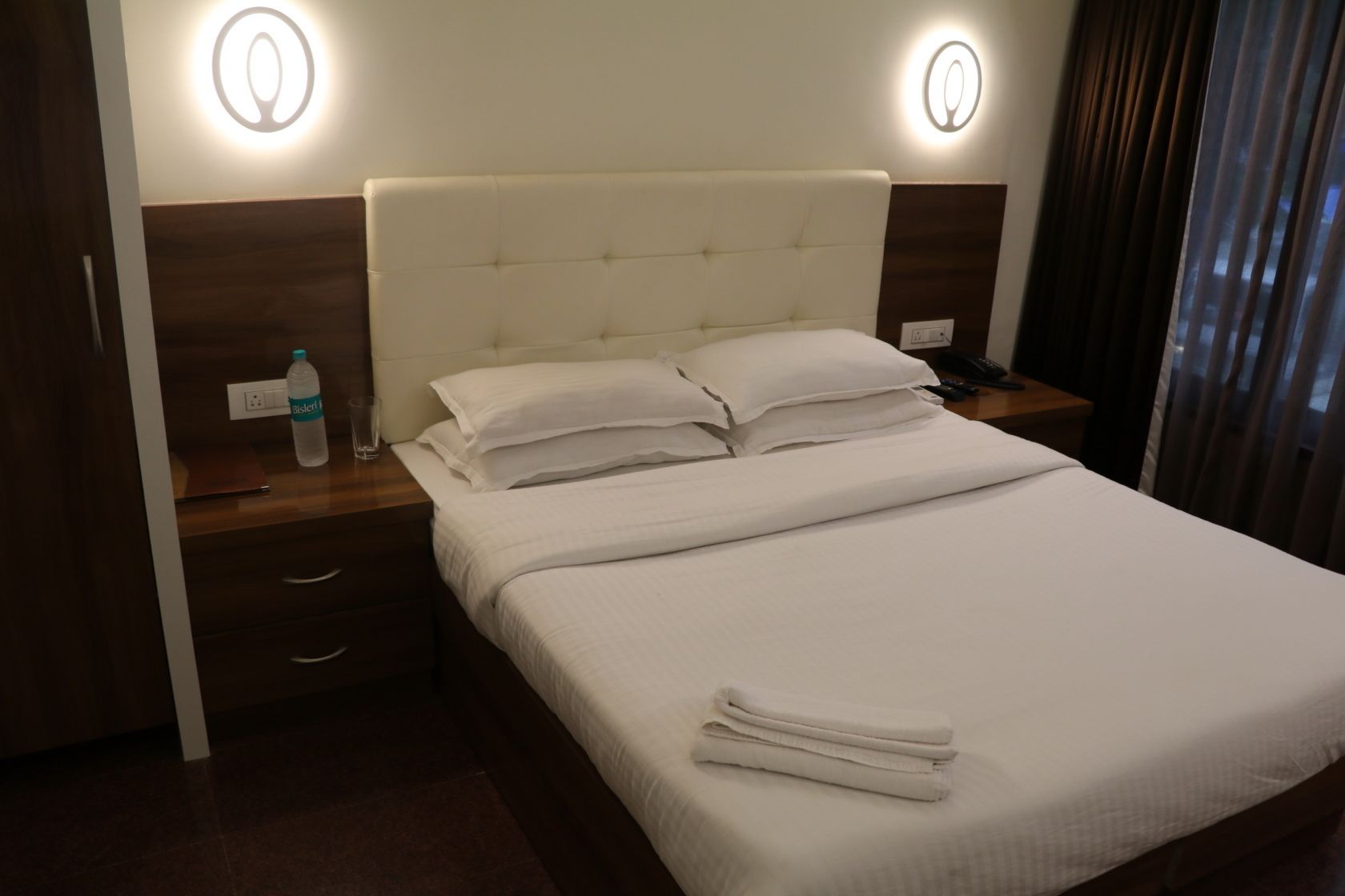 A hotel room with a bed, two bedside tables, a water bottle, a glass, and a phone.