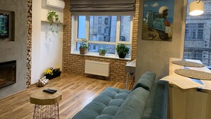 A cozy living room with a sofa, a small round table, a fireplace, and a window with a view of buildings. - KievRent Apartments (Kyiv)