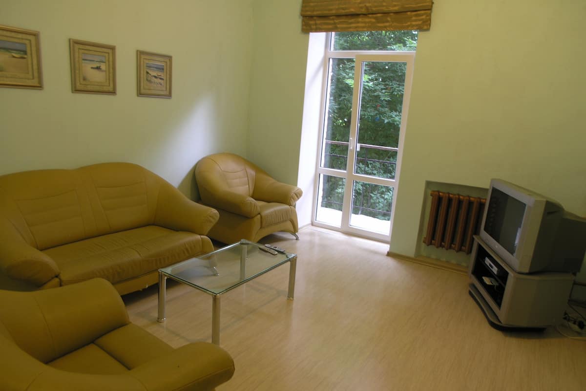A living room with a sofa, armchair, and a glass coffee table. There is a window with a view of trees and a radiator.