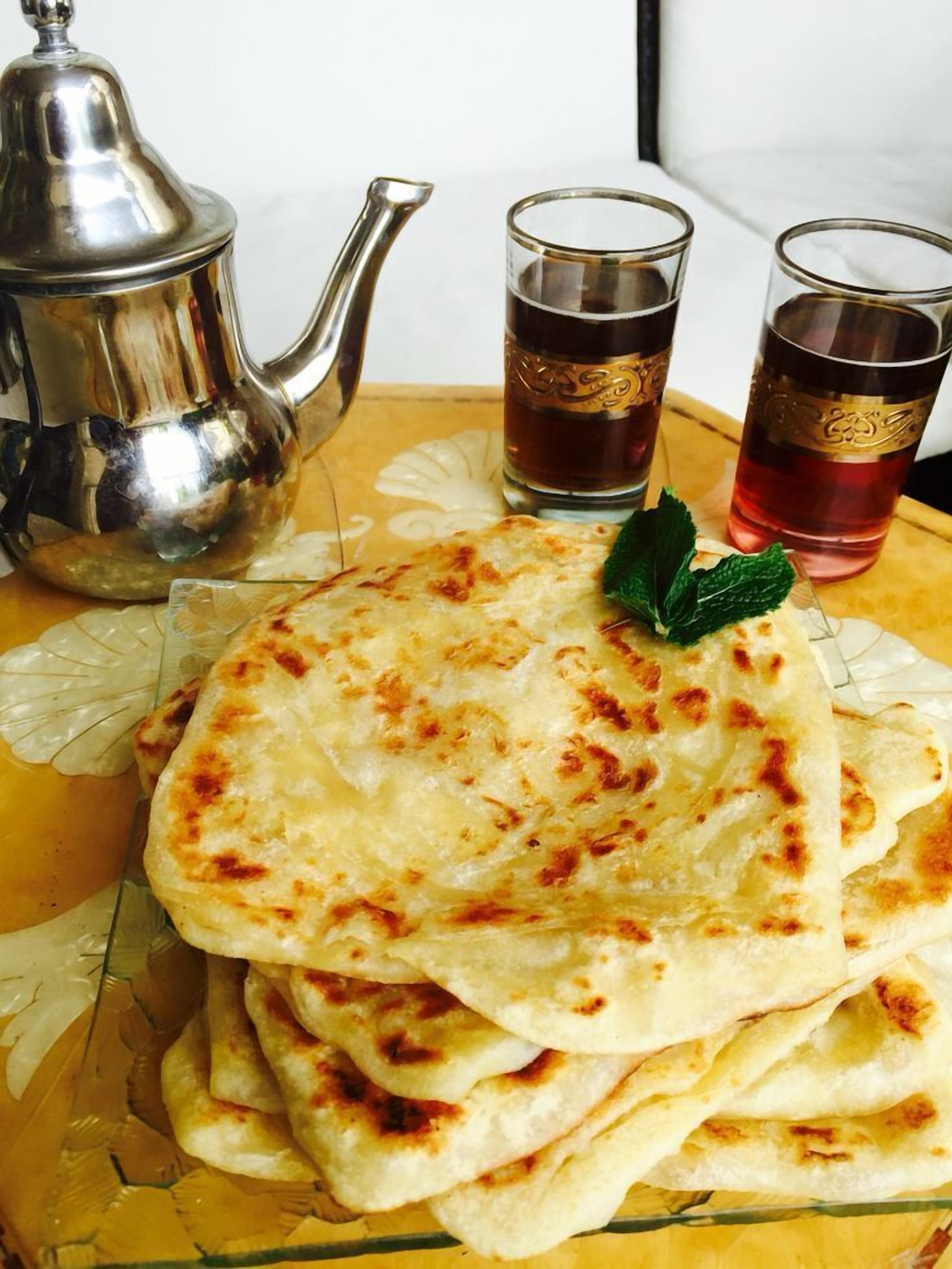 A stack of flatbreads on a glass plate, accompanied by a silver teapot and two glasses of tea.