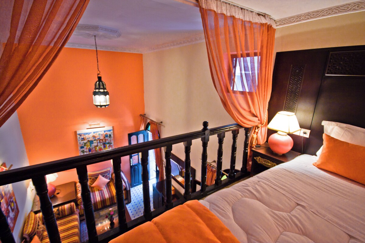 A hotel room with a balcony, a bed with orange bedding, a nightstand with a lamp, and a view of the room below.