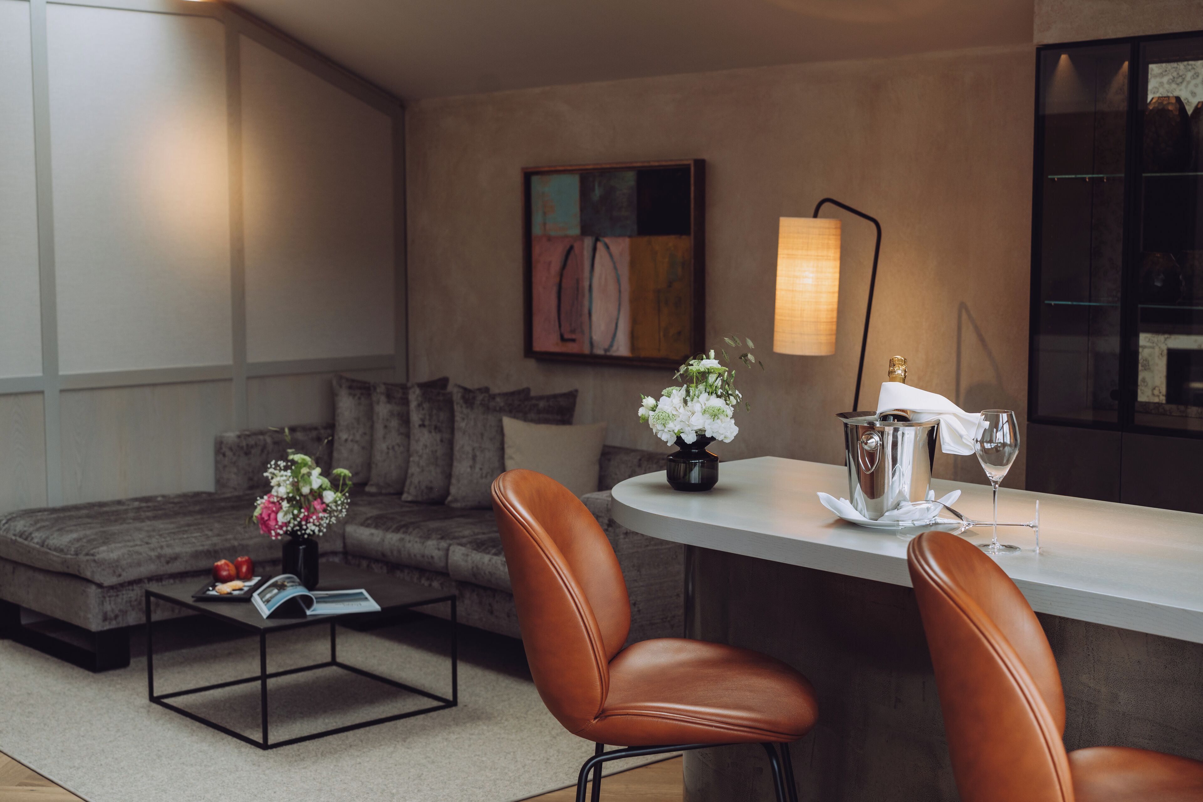 Una sala de estar moderna con un sofá, dos sillones de cuero, una mesa de centro con una revista y flores, una pequeña zona de bar con una copa de vino y una botella, y un cuadro en la pared.
