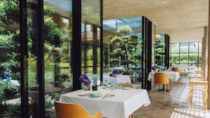 Un restaurant avec des tables dressées pour le service, de grandes fenêtres et une vue sur la verdure environnante.