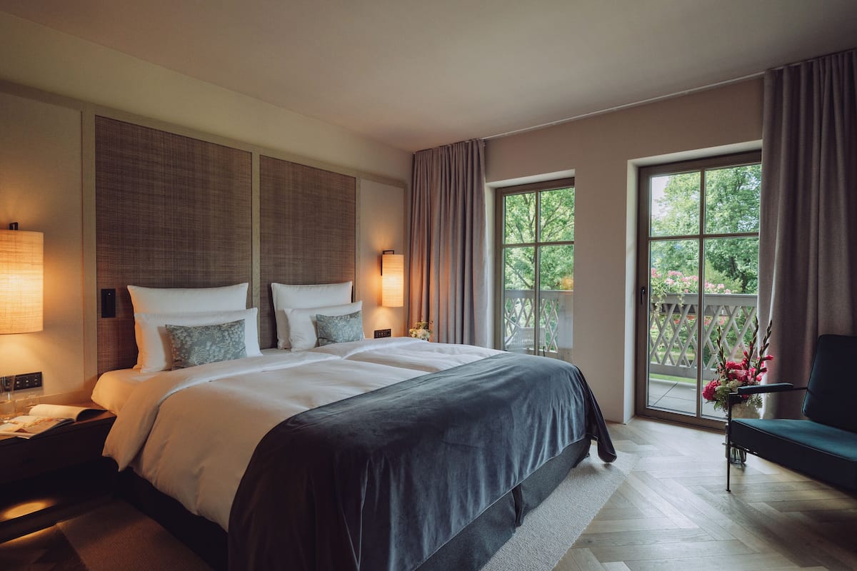 Une chambre d’hôtel moderne avec un grand lit, deux lampes de chevet, une table de chevet, une chaise et une vue sur la verdure par la fenêtre.