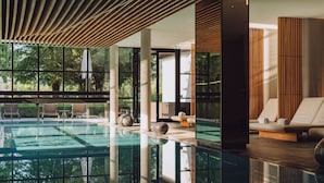 Un espace piscine intérieur moderne, doté de parois en verre transparent, de lambris en bois et de coins salon confortables.