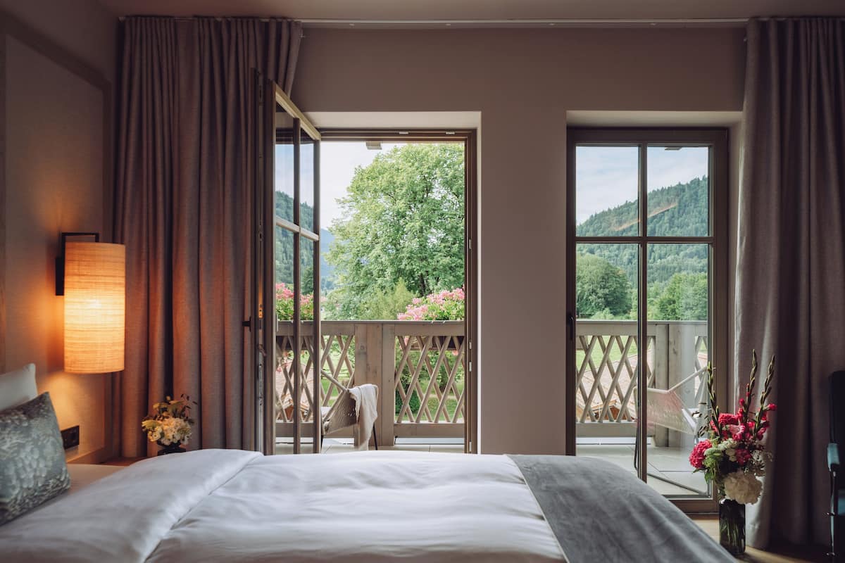 Une chambre d’hôtel avec un grand lit, une table de chevet, une lampe et une vue sur un balcon agrémenté d’une chaise et de fleurs.