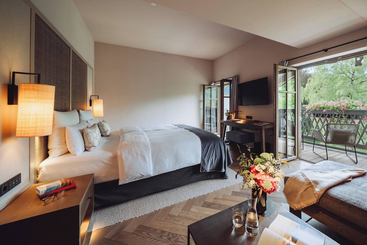 Une chambre d’hôtel moderne dotée d’un grand lit, d’un bureau, d’une télévision et d’un balcon offrant une vue sur les arbres et les fleurs.