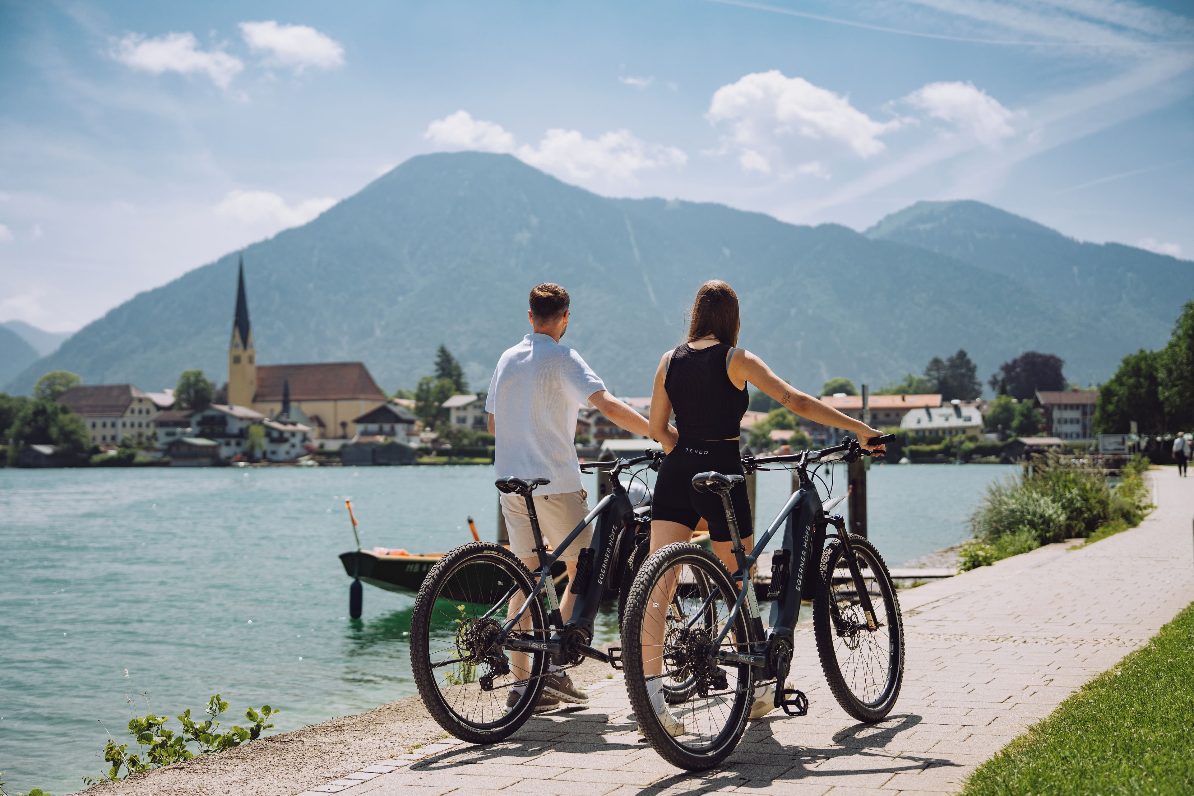 Un couple se tient au bord d’un sentier qui longe un lac, à côté de leurs vélos, et contemple une ville et des montagnes au loin.