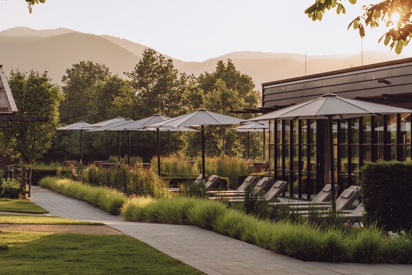 A row of sun loungers with umbrellas, a paved walkway, and a backdrop of trees and mountains. - Parkhotel Egerner Höfe (Rottach-Egern)