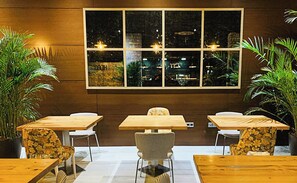 Ein Restaurant mit Holztischen und -stühlen, einem großen Fenster mit Blick auf die beleuchtete Stadt und Blumentöpfen.