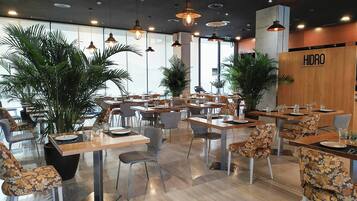 Een restaurantinterieur met tafels, stoelen, hangende lampen en potplanten.