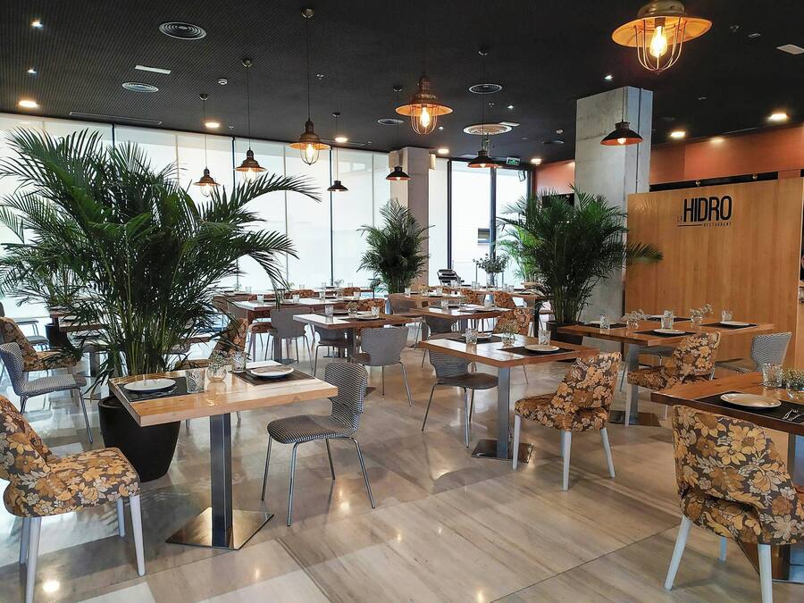 Interior de un restaurante con mesas, sillas, lámparas colgantes y plantas en macetas.
