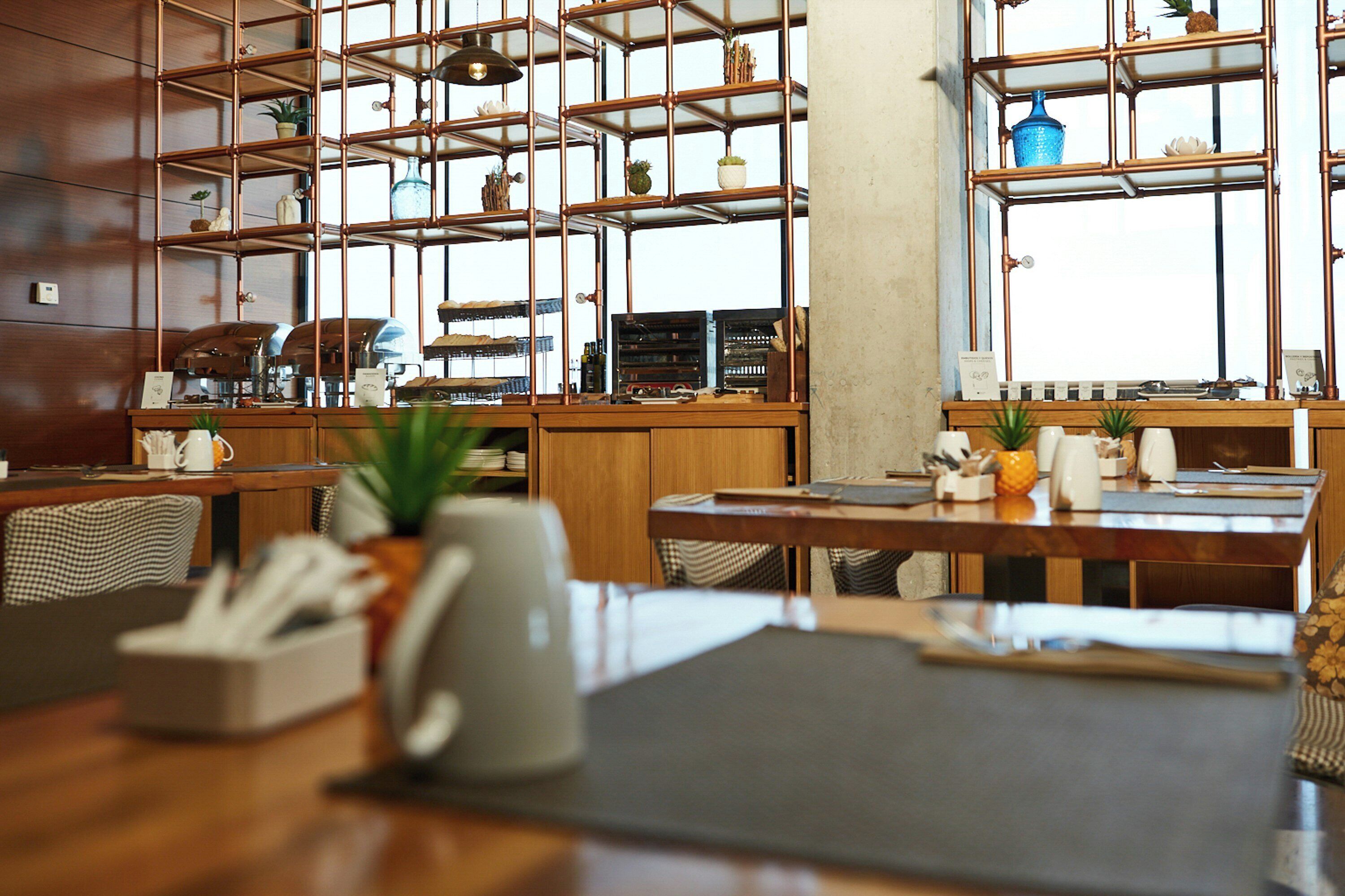 Interior de restaurante con mesas y sillas de madera, y una estantería metálica con plantas.
