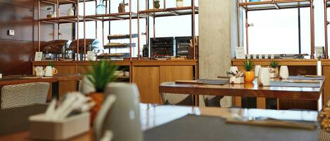 Interior de restaurante con mesas y sillas de madera, y una estantería metálica con plantas.