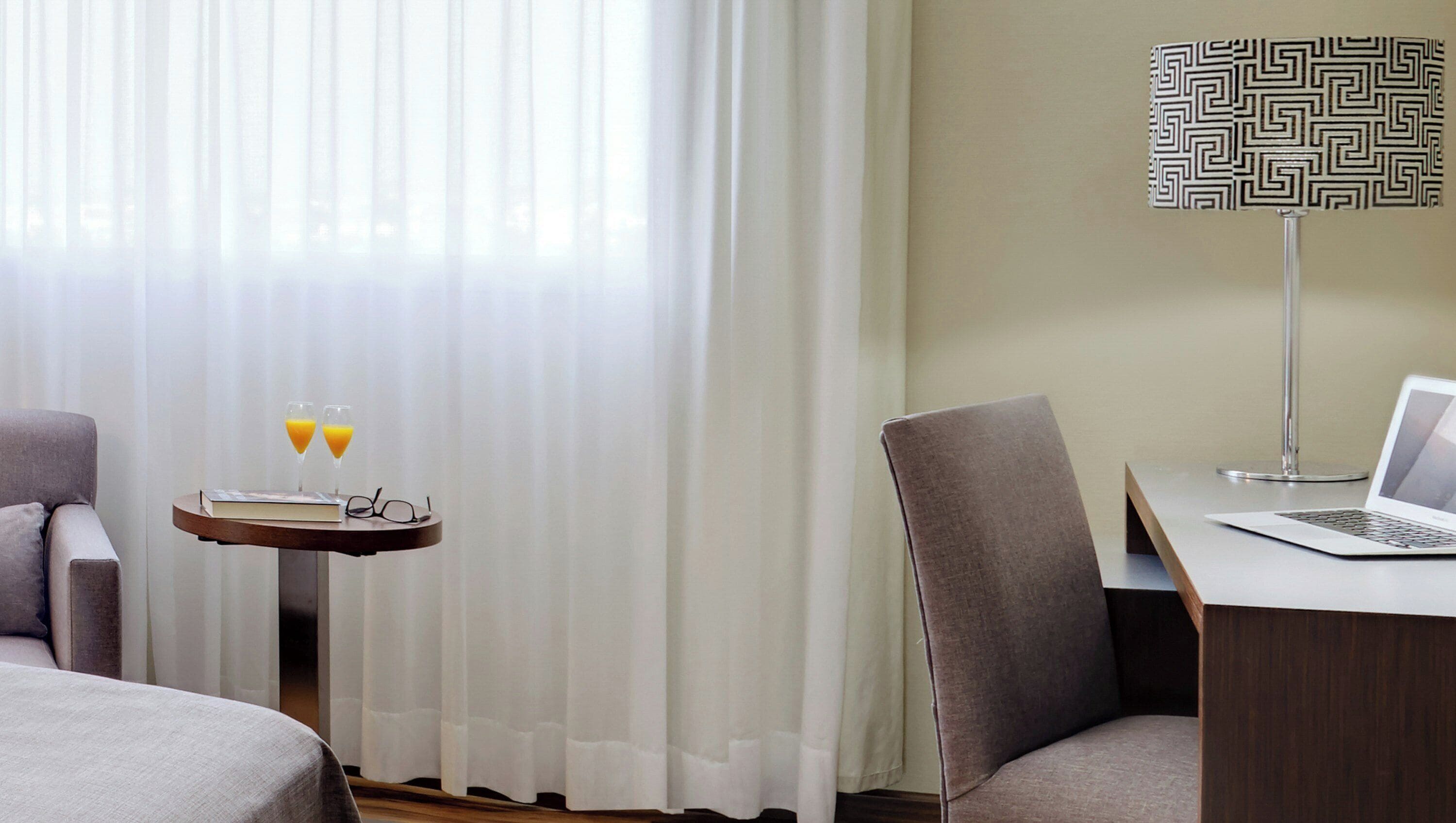 A hotel room with a bed, a desk with a laptop, a chair, a bedside table with glasses and juice glasses, and a lamp with a geometric pattern.
