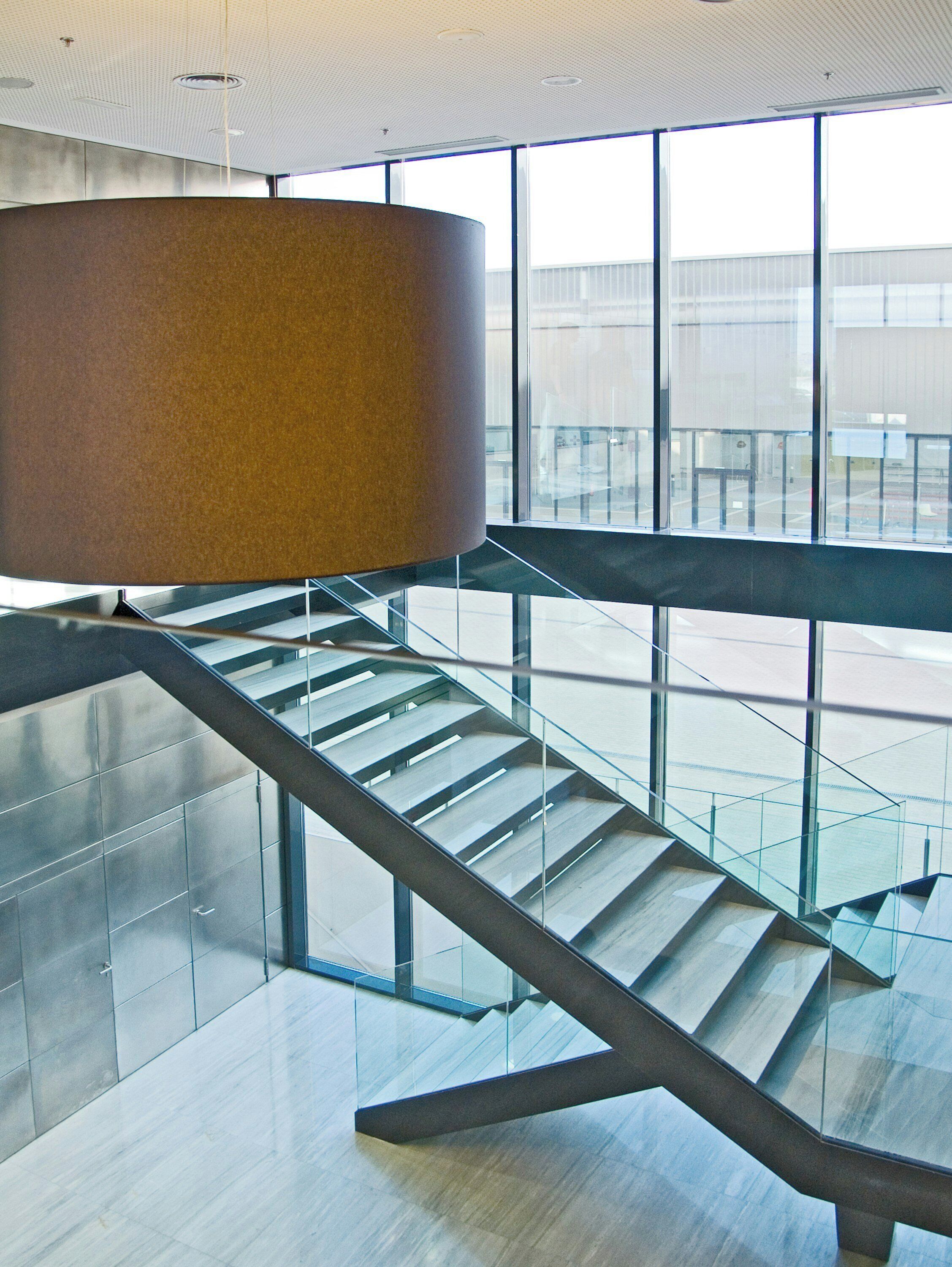 Un intérieur moderne avec un escalier en verre, un grand luminaire cylindrique et une vue dégagée sur l’extérieur grâce à de grandes fenêtres.