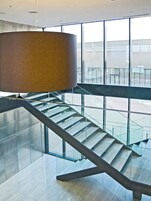 Un interior moderno con una escalera de cristal, una gran lámpara cilíndrica y vistas amplias por ventanas grandes.
