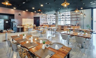 A modern dining area with multiple tables and chairs, a kitchen in the background, and pendant lighting.